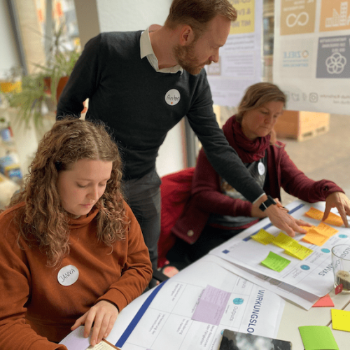 2 Personen sitzen eine Person steht dahinter. Alle schauen auf den Tisch und Arbeiten am Projekt.